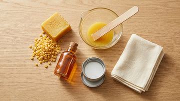 Overhead flat-lay of DIY leather conditioner ingredients including beeswax, neatsfoot oil, glass bowl, and metal tin on a wooden surface