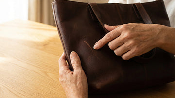 Hands examining a surface scratch on a dark brown genuine leather bag