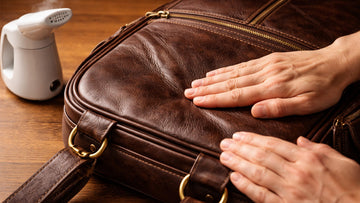 Hands smoothing a crease on a dark brown genuine leather bag after steaming on a wooden surface