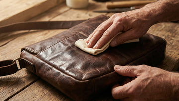 Hands applying leather conditioner to a dark brown leather bag on a wooden workbench