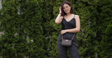 Stylish model in black top and glasses, subtly posing with a patterned designer handbag against a green hedge. Ideal for accessory e-commerce photos.