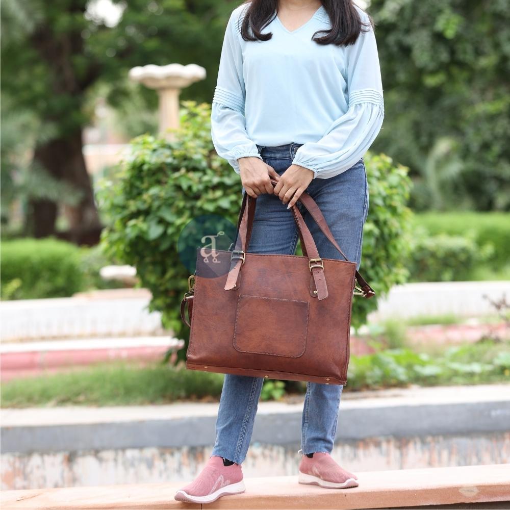 Woman Standing Outdoors Holding a Brown Leather Laptop Tote Bag with Brass Buckles and Front Pocket, Wearing Casual Outfit