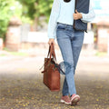 Woman Walking Outdoors Carrying Brown Leather Laptop Tote Bag with Brass Buckles, Wearing Casual Jeans and Sneakers