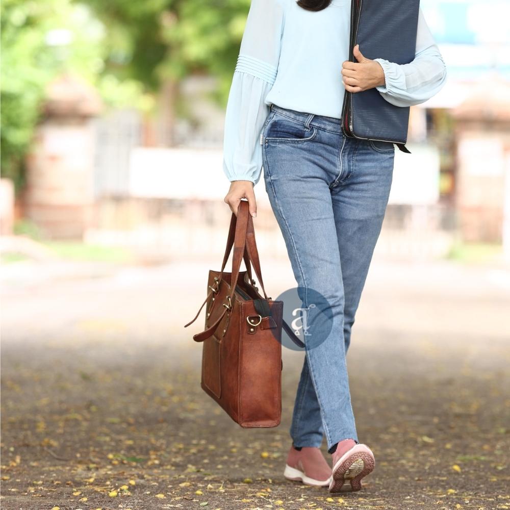 Woman Walking Outdoors Carrying Brown Leather Laptop Tote Bag with Brass Buckles, Wearing Casual Jeans and Sneakers
