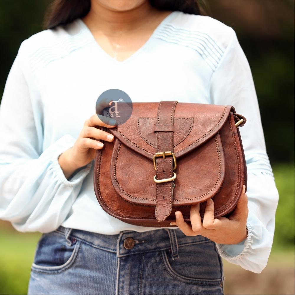 Close-Up of Woman Holding a Brown Leather Saddle Purse with Buckle, Dressed in Light Blue Blouse and Denim Jeans