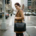Woman in a camel coat at a city crossing wearing The Virtuoso black leather briefcase on its shoulder strap