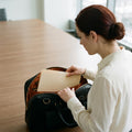 Woman at a desk sliding a paper folder into the open main compartment of The Virtuoso black leather briefcase