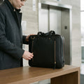 Woman by a lift opening a back zip pocket of The Virtuoso black leather briefcase standing on a counter