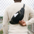 Man wearing a black leather fanny pack across his back, reaching into its zip pocket
