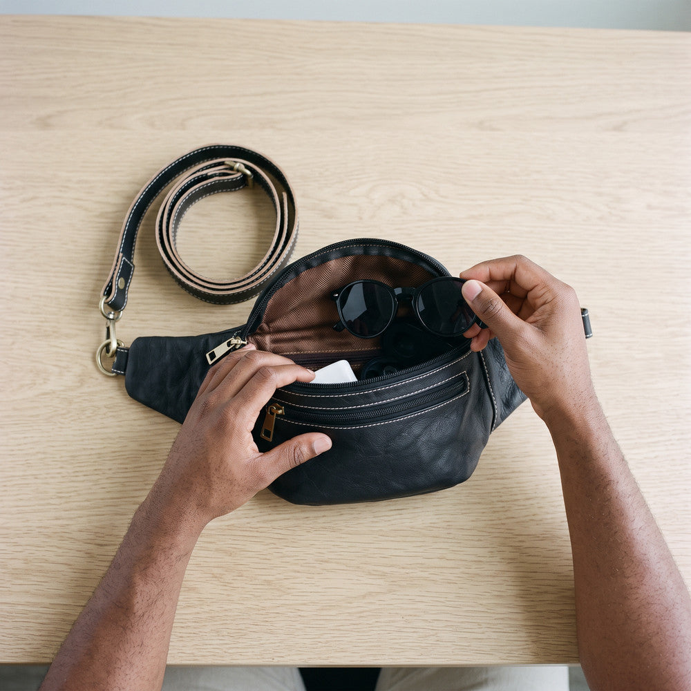 Hands opening a black leather fanny pack on a wooden table, with sunglasses inside