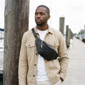Man in a beige jacket wearing a black leather fanny pack across his chest on a harbour boardwalk