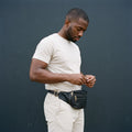 Man in a cream T-shirt with a black leather fanny pack worn at the waist against a dark wall
