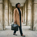 Woman in a camel coat walking through a stone colonnade carrying The Specialist black leather laptop briefcase by its handles