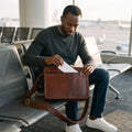Man at an airport gate sliding a boarding pass into the back zip pocket of The Postman leather messenger bag