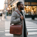 Man at a city crosswalk adjusting the shoulder strap of The Postman leather computer messenger bag