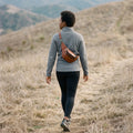 Woman in a grey fleece walking up a hillside trail with the brown leather fanny pack slung across her back