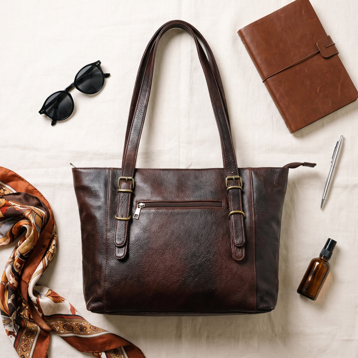 Flat lay of the dark brown leather purse with a silk scarf, notebook, sunglasses, pen and perfume