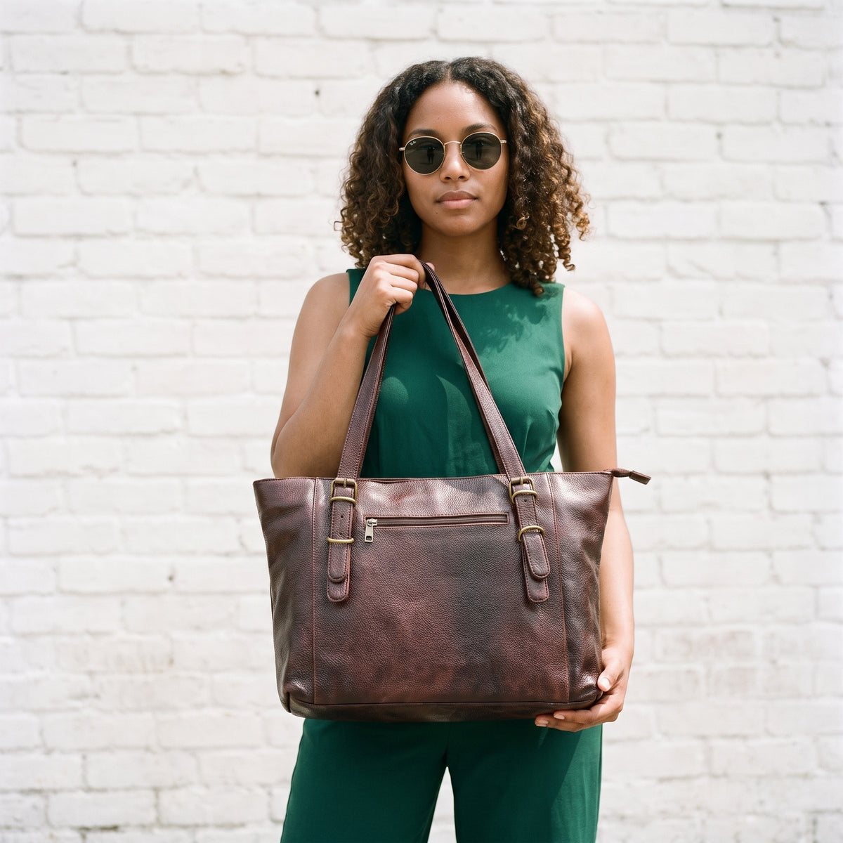 Woman in a green jumpsuit and sunglasses holding the dark brown leather purse on her shoulder by a white brick wall