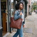 Woman in a denim jacket walking past a bookshop wearing the Brown Leather Tote Bag on her shoulder