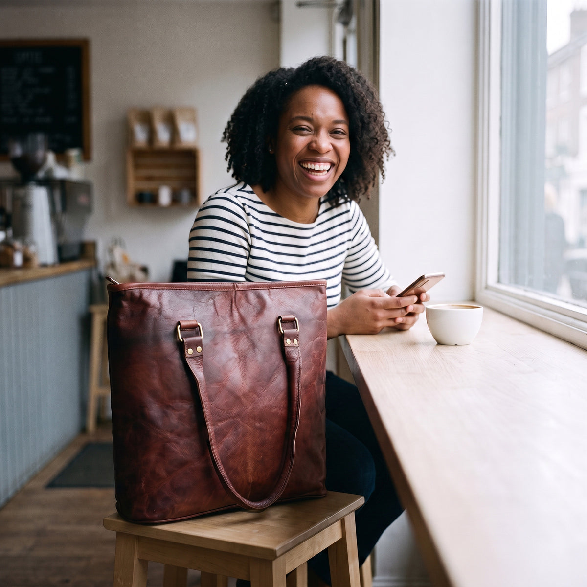 Smiling woman at a cafe window counter with the Brown Leather Tote Bag on the stool beside her