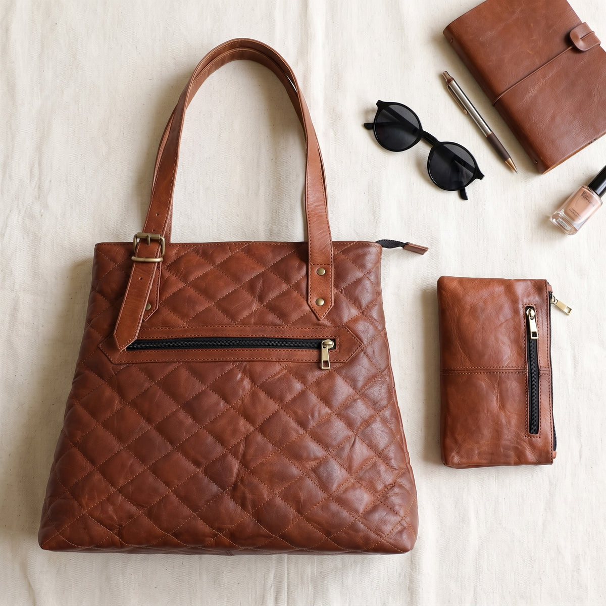 Flat lay of the quilted brown leather designer tote bag with a matching pouch, notebook, pen and sunglasses