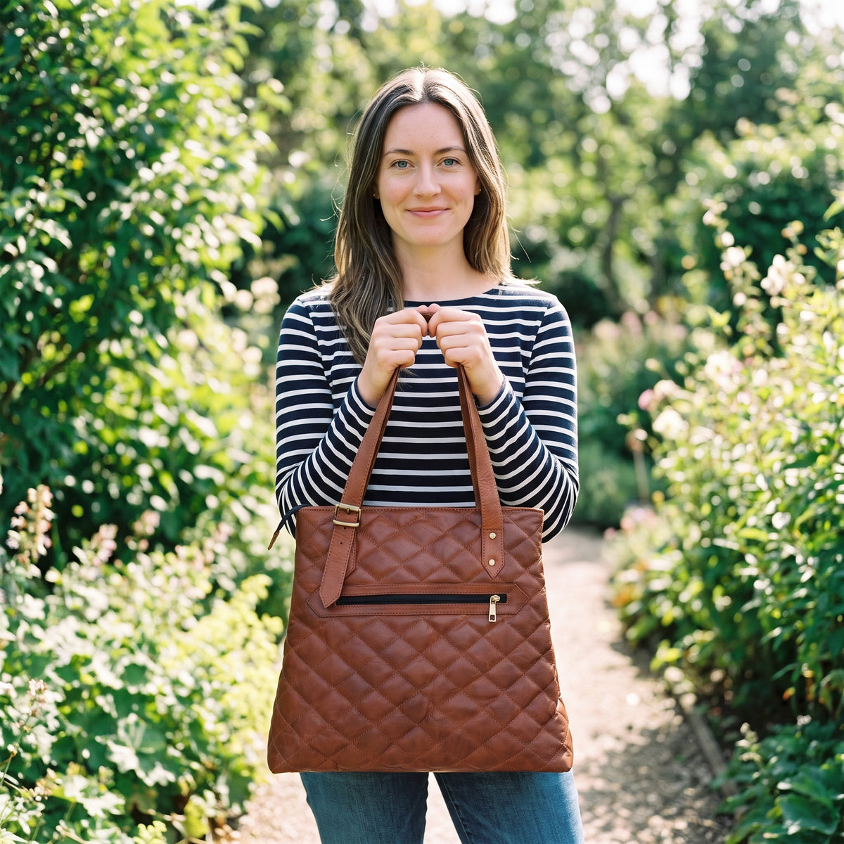 Woman in a striped top holding the quilted brown leather designer tote bag by its handles on a garden path