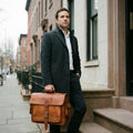 Man in a charcoal overcoat holding The Executive tan leather computer briefcase by its top handle on a townhouse doorstep