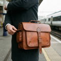 Hand opening the side pocket of The Executive tan leather briefcase on a train station platform