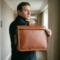 Man holding The Executive tan leather briefcase by its top handle to show the zippered back pocket
