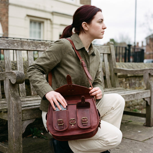 Woman crouched by a park bench lifting the flap of the oxblood leather saddle bag to reach inside