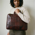 Woman in a cream jumper holding The Negotiator dark brown leather laptop briefcase against her chest by its cut-out handle