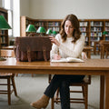 Woman writing at a library table with a hand on the flap of The Digital leather laptop messenger bag