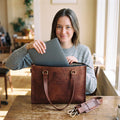 Woman at a cafe table sliding a laptop into the zippered top of the brown leather laptop tote bag, detachable strap beside it
