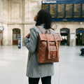 Back view of a brown leather roll-top backpack with buckle straps worn in a railway station hall