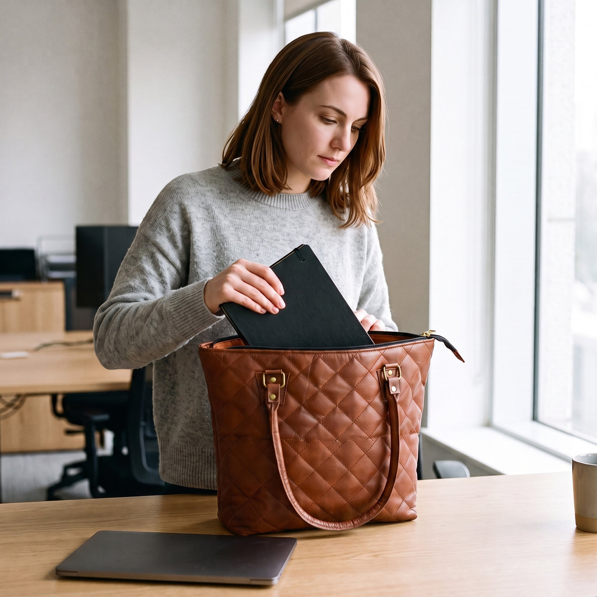 Woman at an office desk sliding a notebook into the open zip top of the quilted Leather Tote Bag for Work